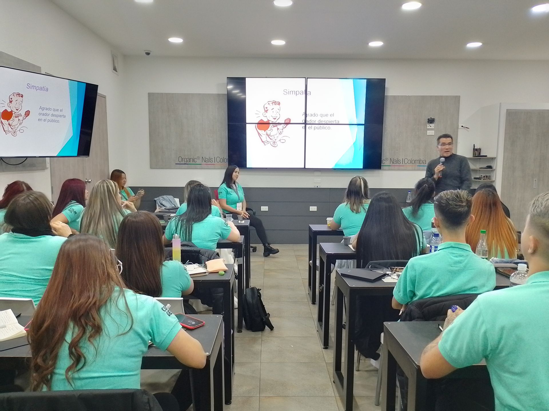 Una presentación en el aula con estudiantes con camisetas verde azulado, el instructor hablando y diagramas en pantallas.