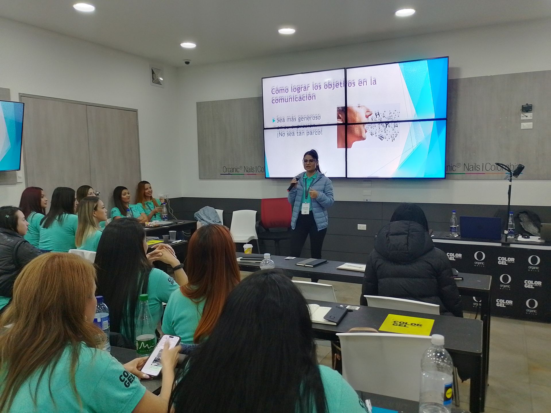 Mujer haciendo una presentación ante un grupo en un aula con una pantalla que muestra texto.