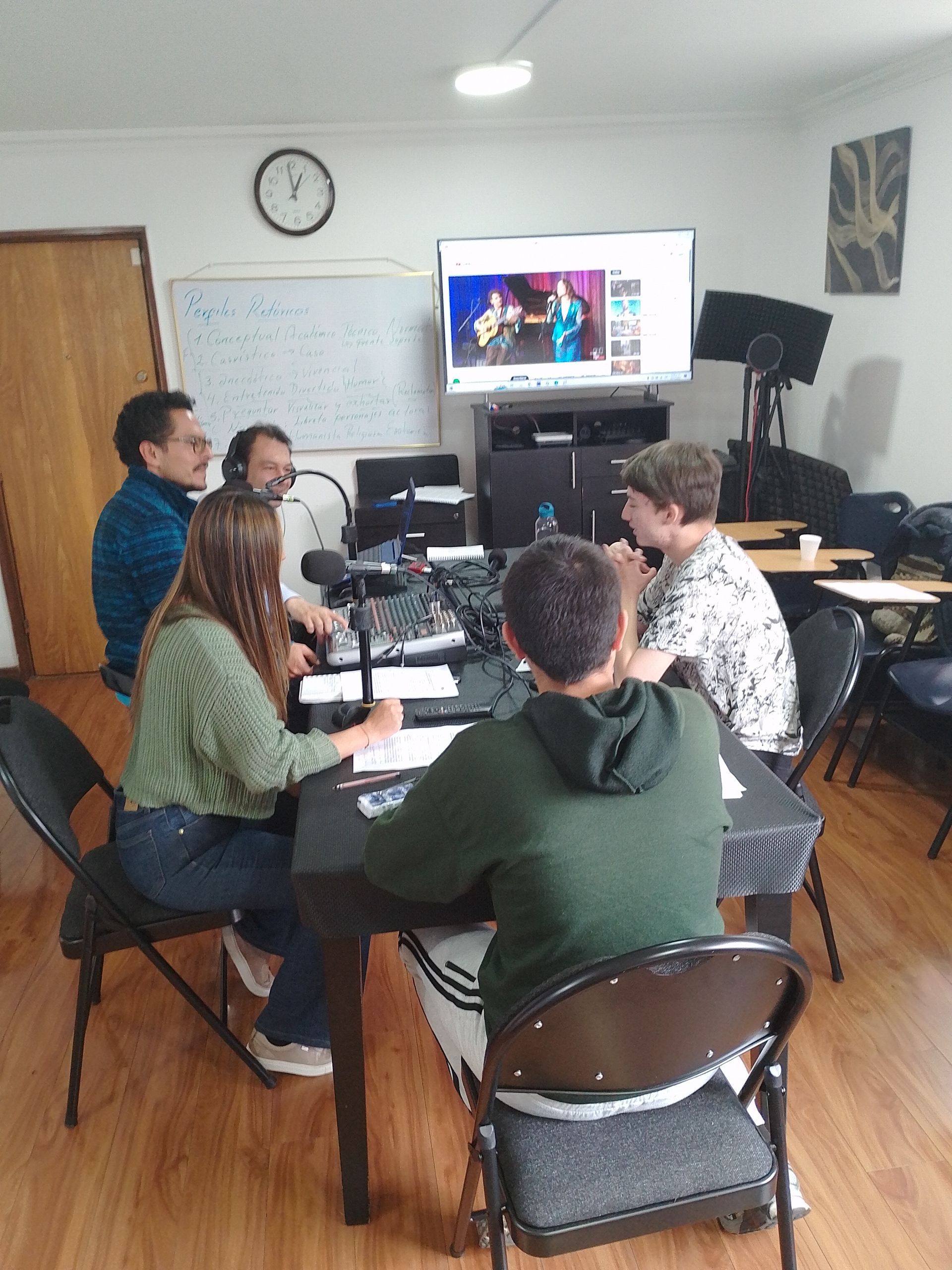 Personas en un estudio, sentadas en una mesa con un micrófono y mirando una pantalla.
