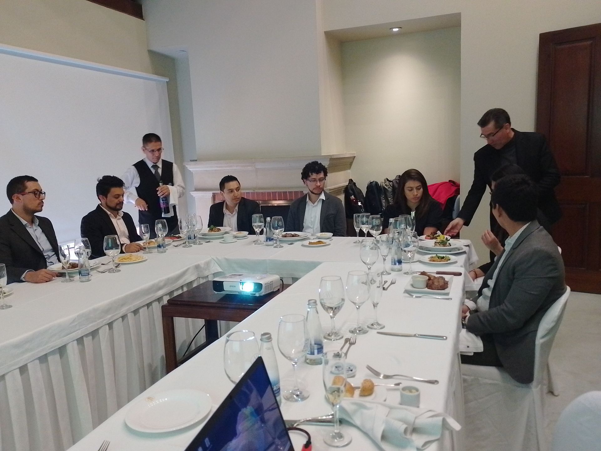 Personas sentadas en una mesa en forma de U, atendidas por camareros, durante un evento formal.