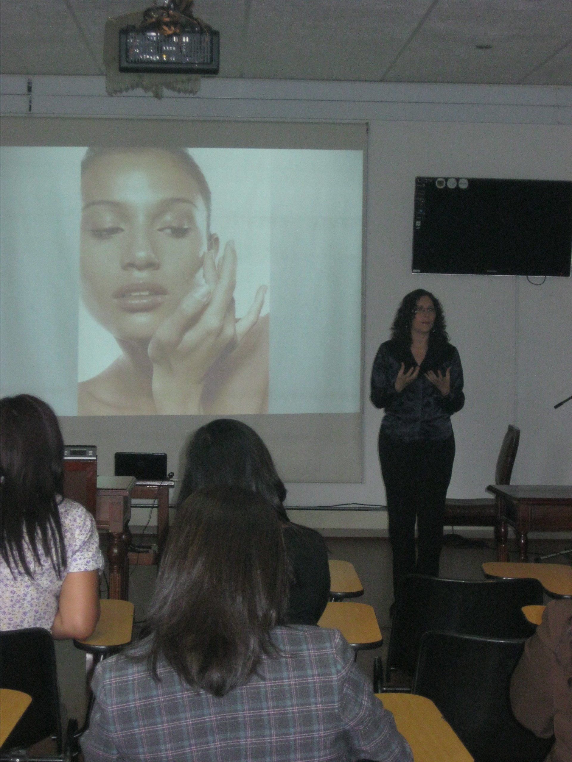 Una mujer presenta en un aula con la imagen de un rostro proyectado, gesticulando hacia él. El público, sentado, observa.