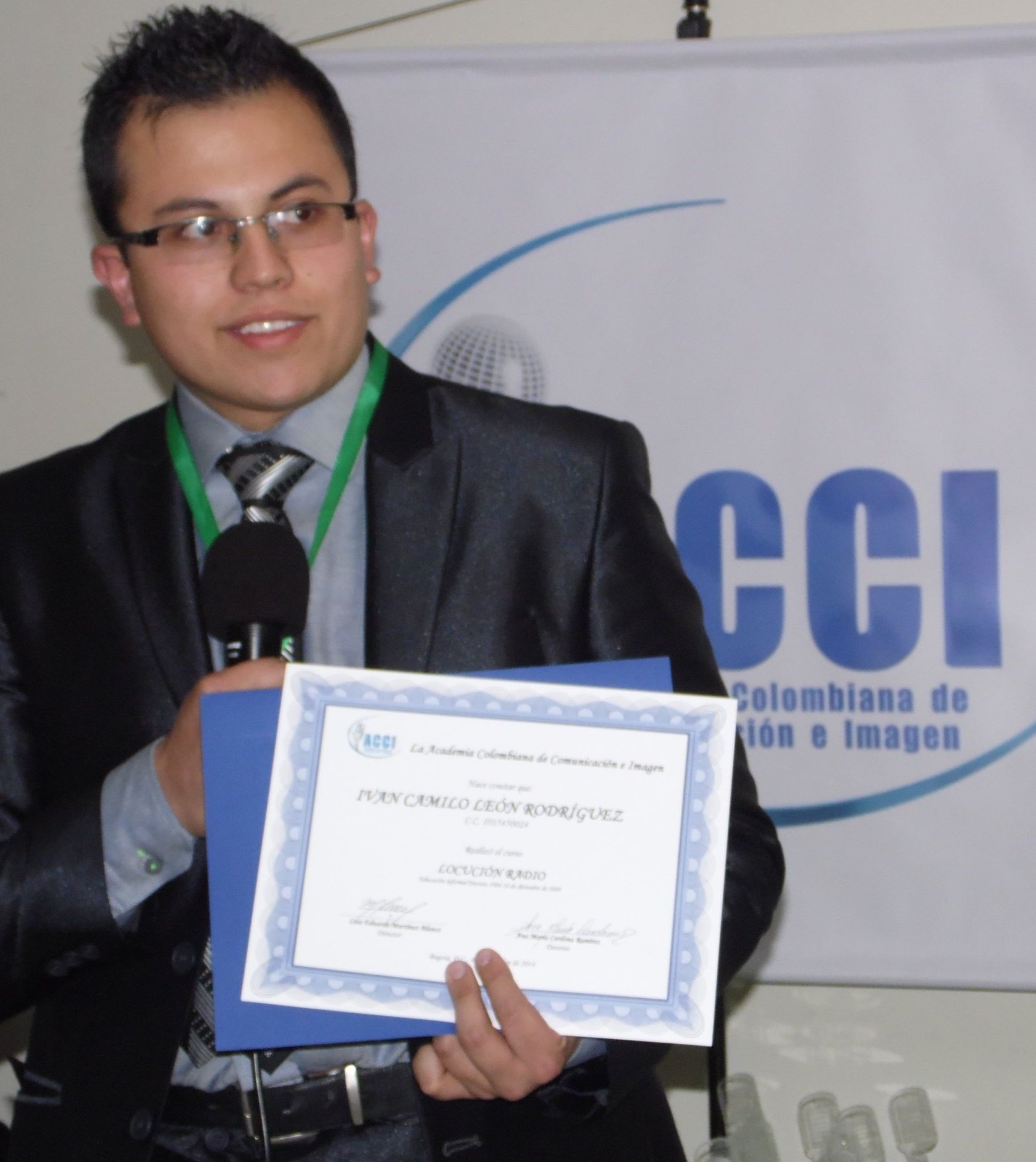 Hombre de traje con certificado, sonriendo y hablando por el micrófono. Fondo azul y blanco con logotipo.