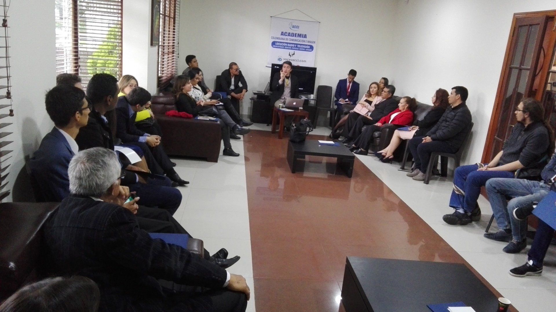 Un grupo de personas sentadas en una habitación, escuchando a una persona en el centro, posiblemente una presentación.