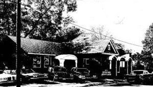 Black-and-white photo of a small building with parked cars and trees in front