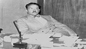 Man seated at a desk with papers, wearing a light suit, in front of wood paneling.