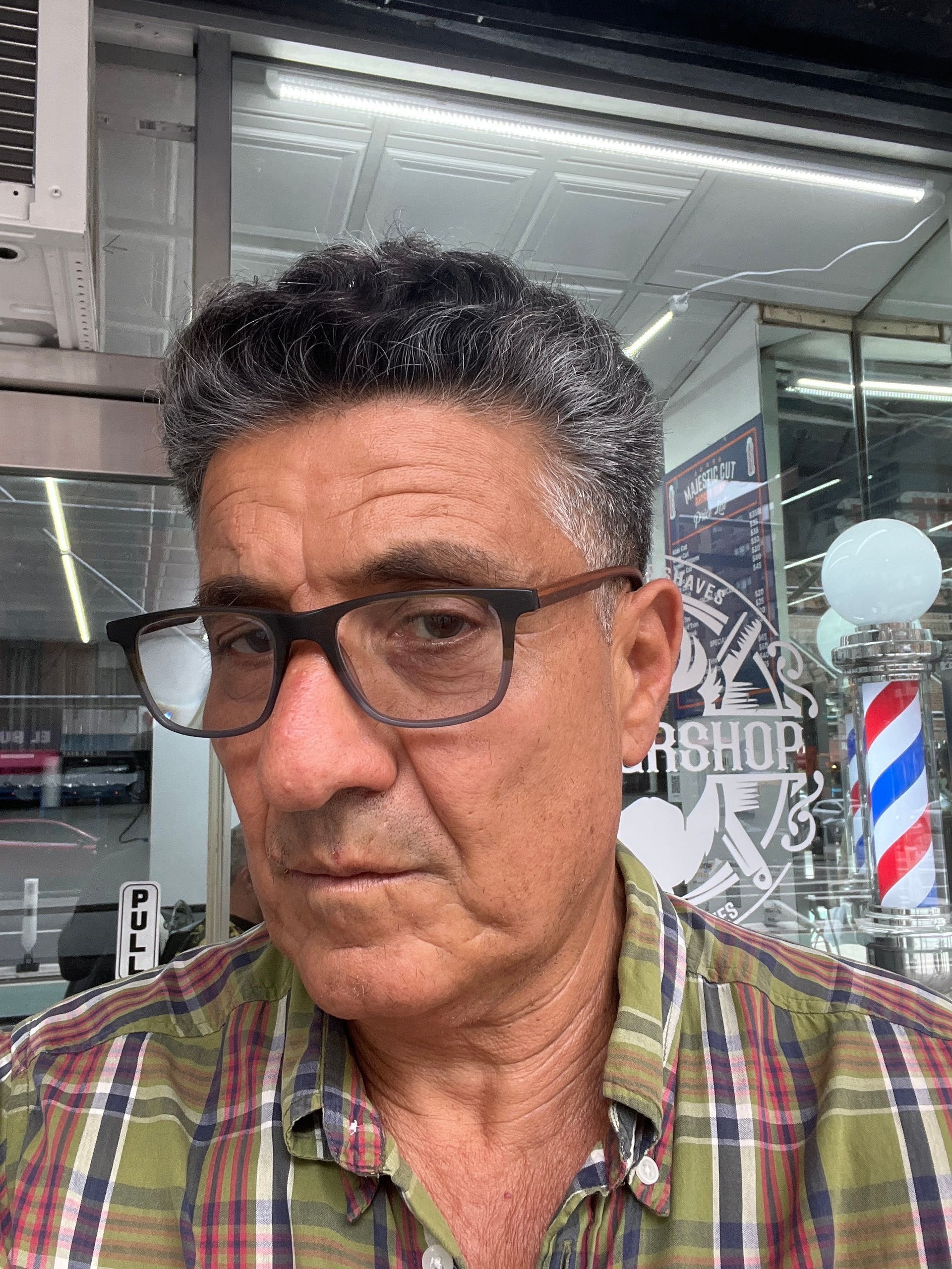 Man wearing glasses in front of a barber shop window, with a red-and-blue barber pole visible.