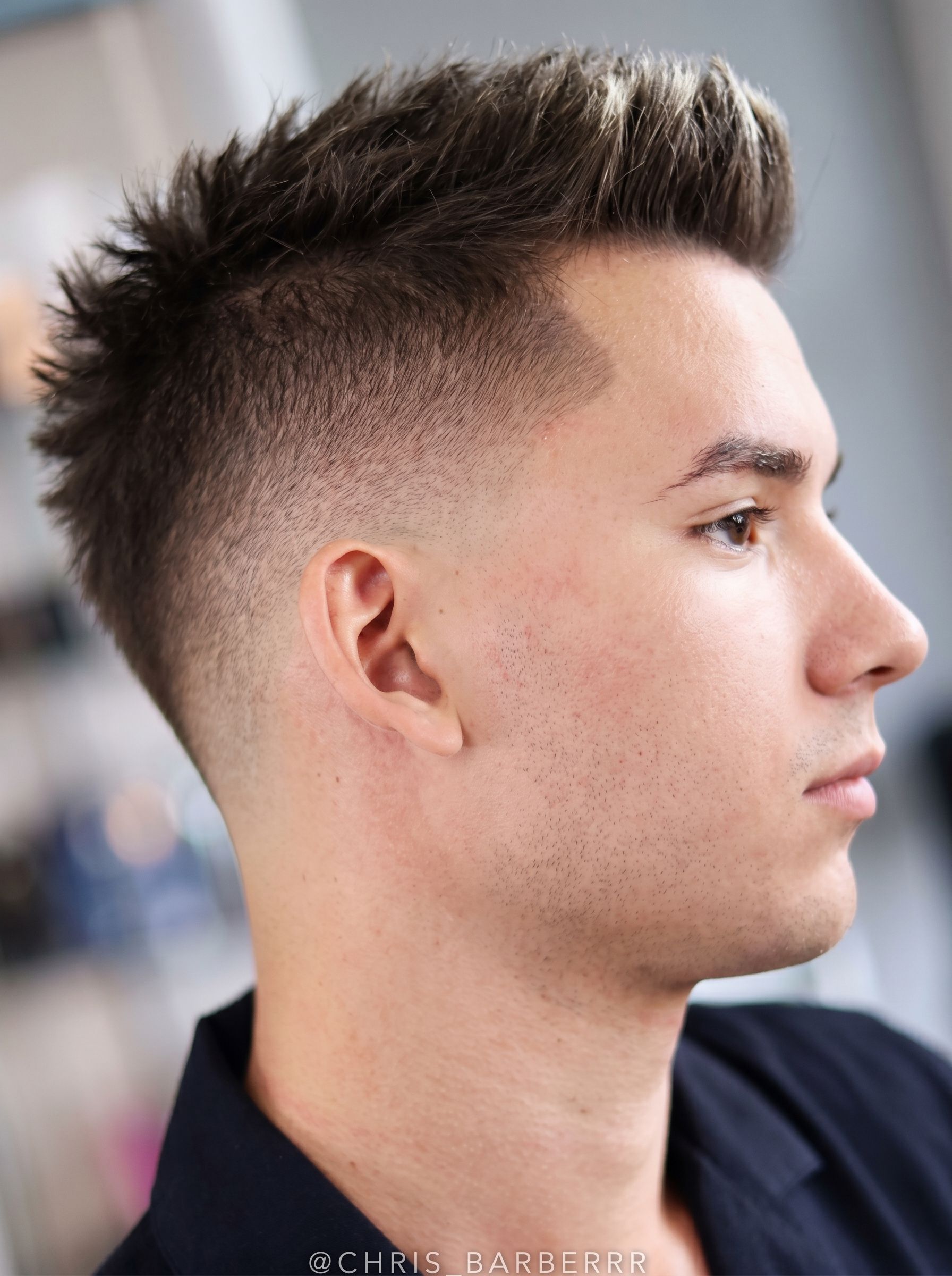 Profile of a man with a short spiky haircut, wearing a dark jacket, against a blurred indoor background