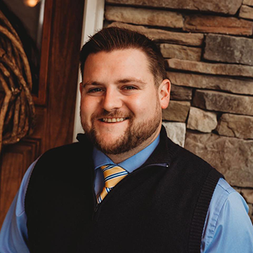 A man in a blue shirt and tie is smiling in front of a stone wall.