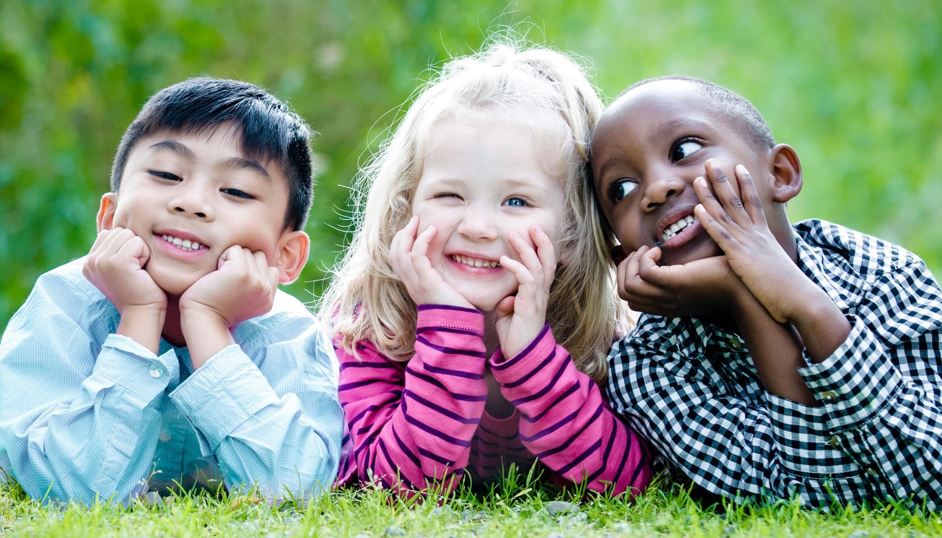 Three children lying on grass, resting their chins in their hands and smiling at the camera.
