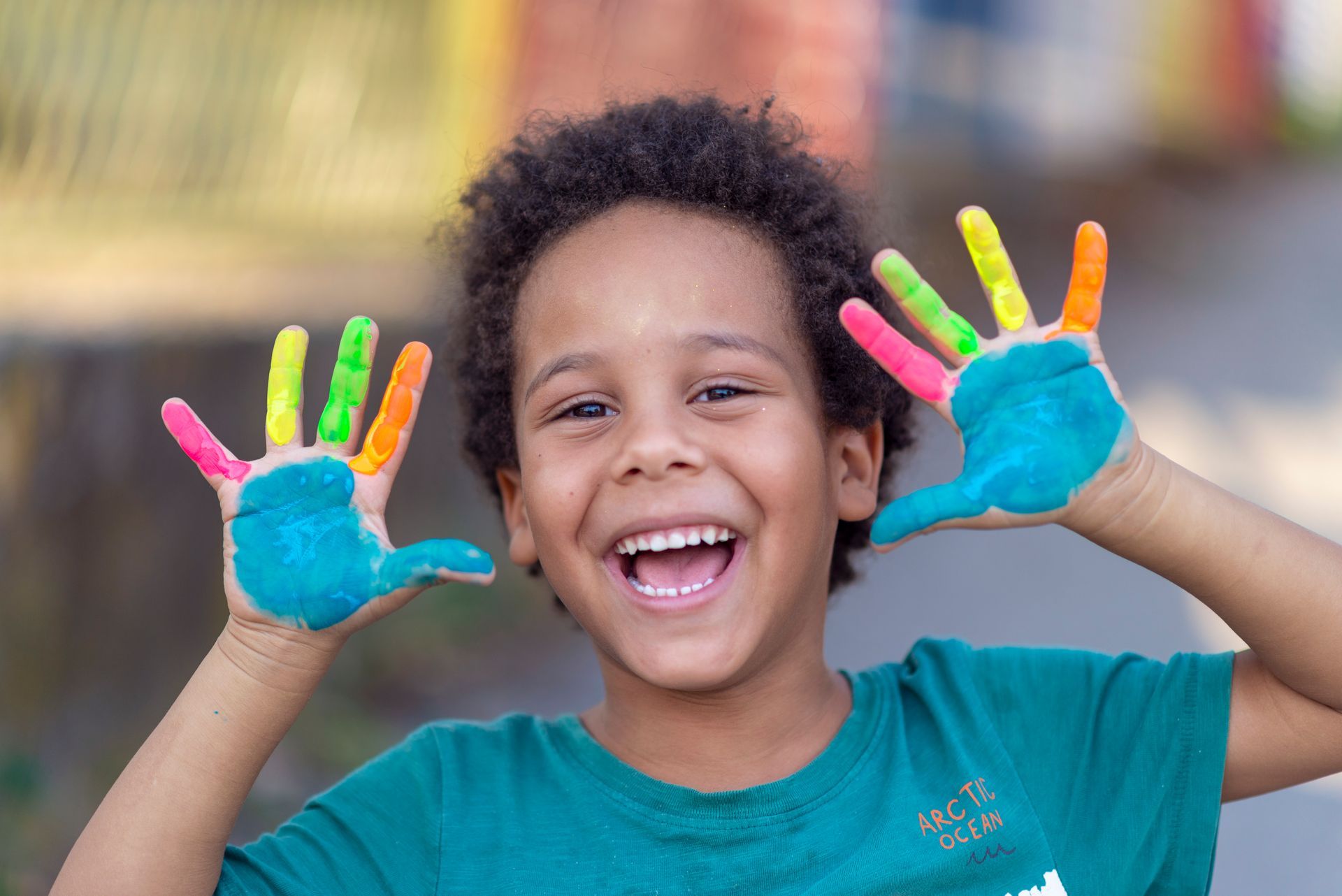 Smiling child with painted hands raised outdoors, wearing a teal shirt.