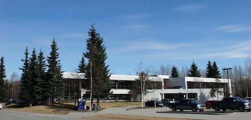 Low building and parking lot with evergreen trees under a clear blue sky