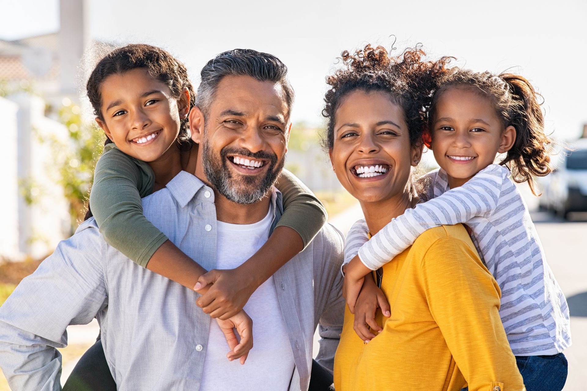 Smiling family of four outdoors, two children hugging adults in bright sunlight.