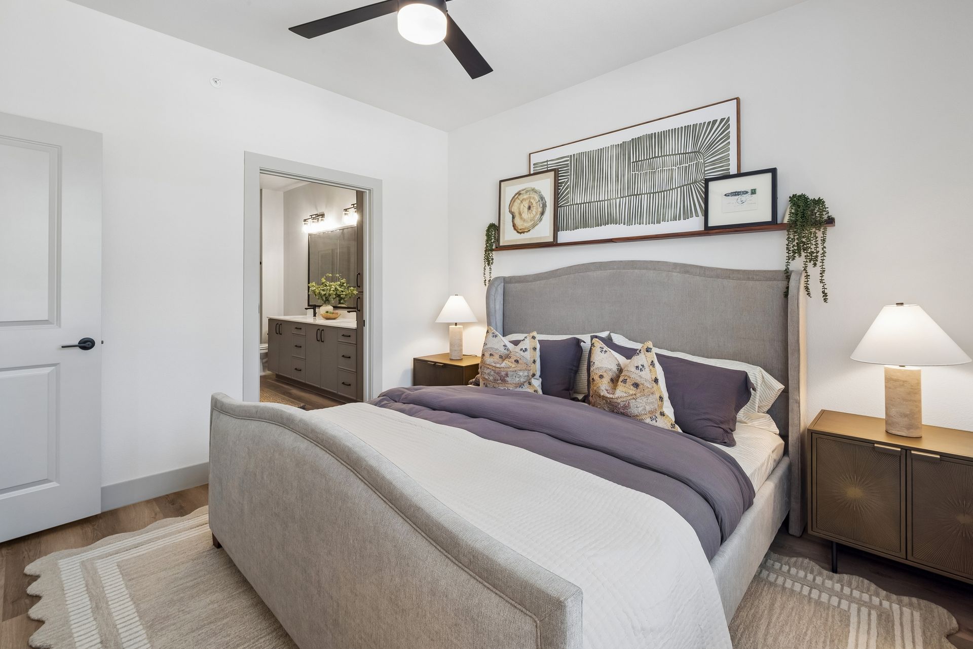 Bright bedroom with a gray upholstered bed, white walls, wooden nightstands, and a ceiling fan.