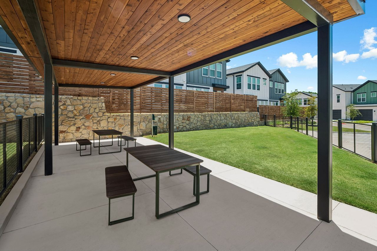 Covered patio with outdoor dining table, benches, and grassy backyard beside modern homes