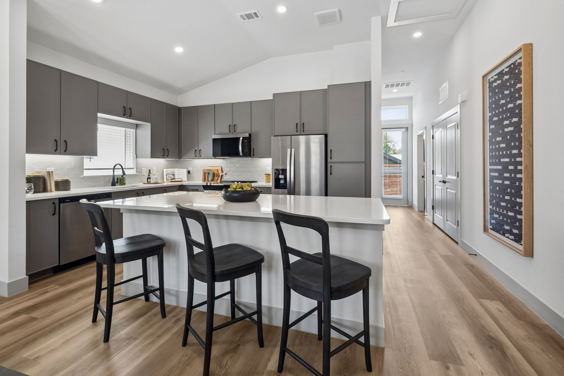 Modern kitchen with gray cabinets, white island, three black stools, and stainless steel appliances.