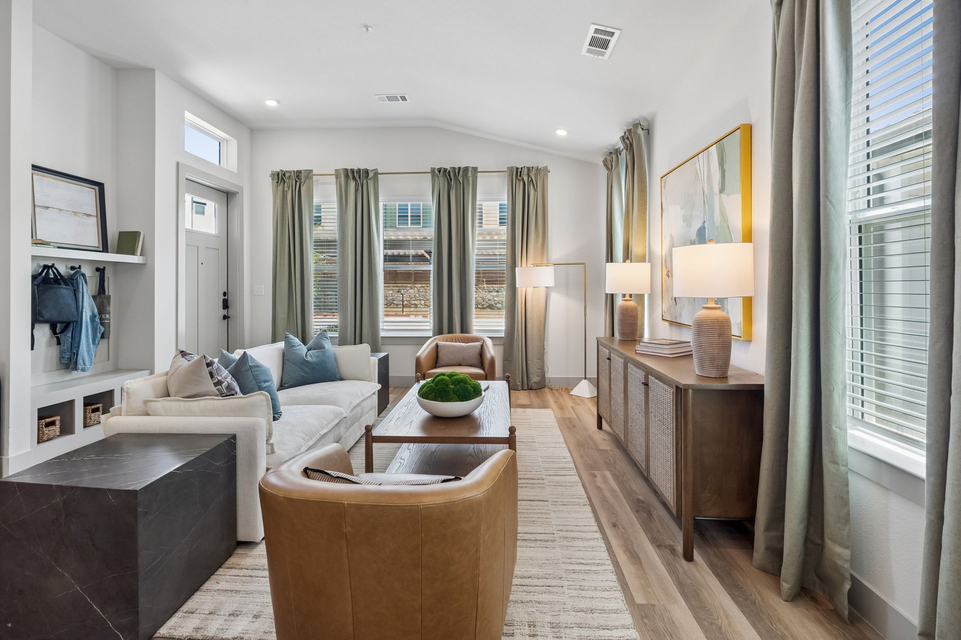 Bright modern living room with sofa, armchair, sideboard, lamps, and large windows