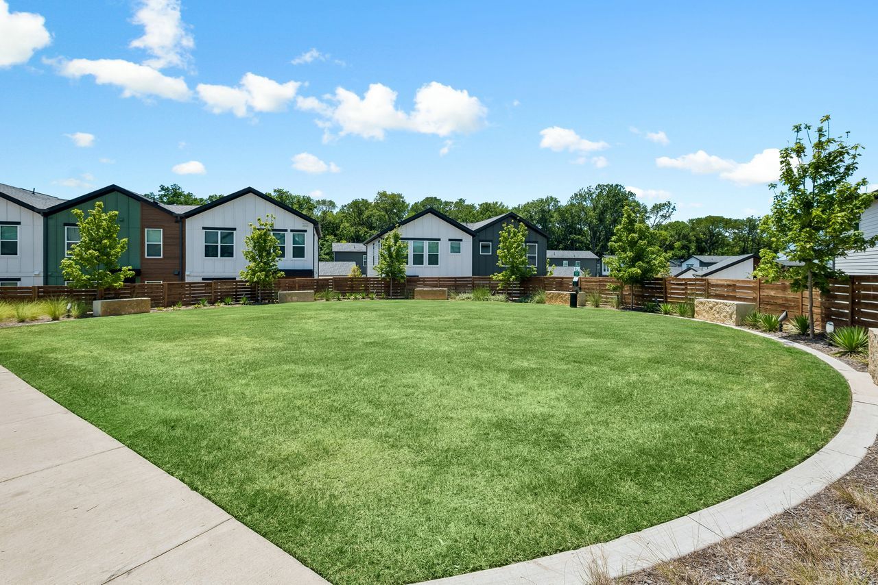 Curved grassy common area with sidewalk, fence, and row of modern houses under a blue sky