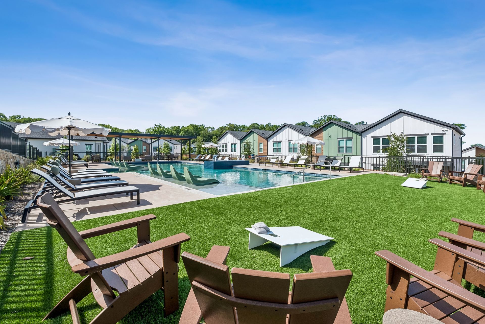 Poolside lounge area with green lawn, chairs, umbrellas, and modern houses in the background