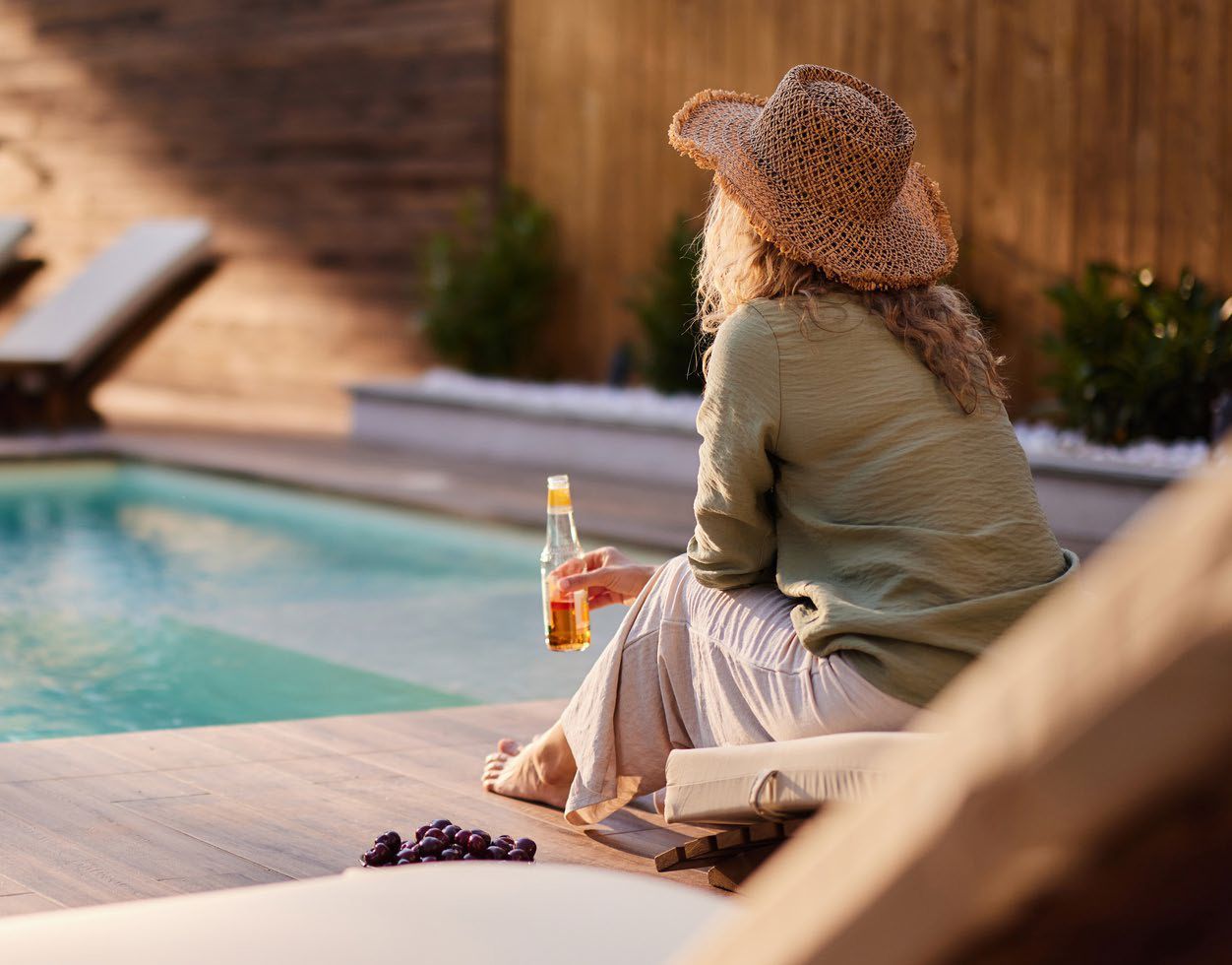 A woman is sitting on a chair by a pool holding a bottle of beer.