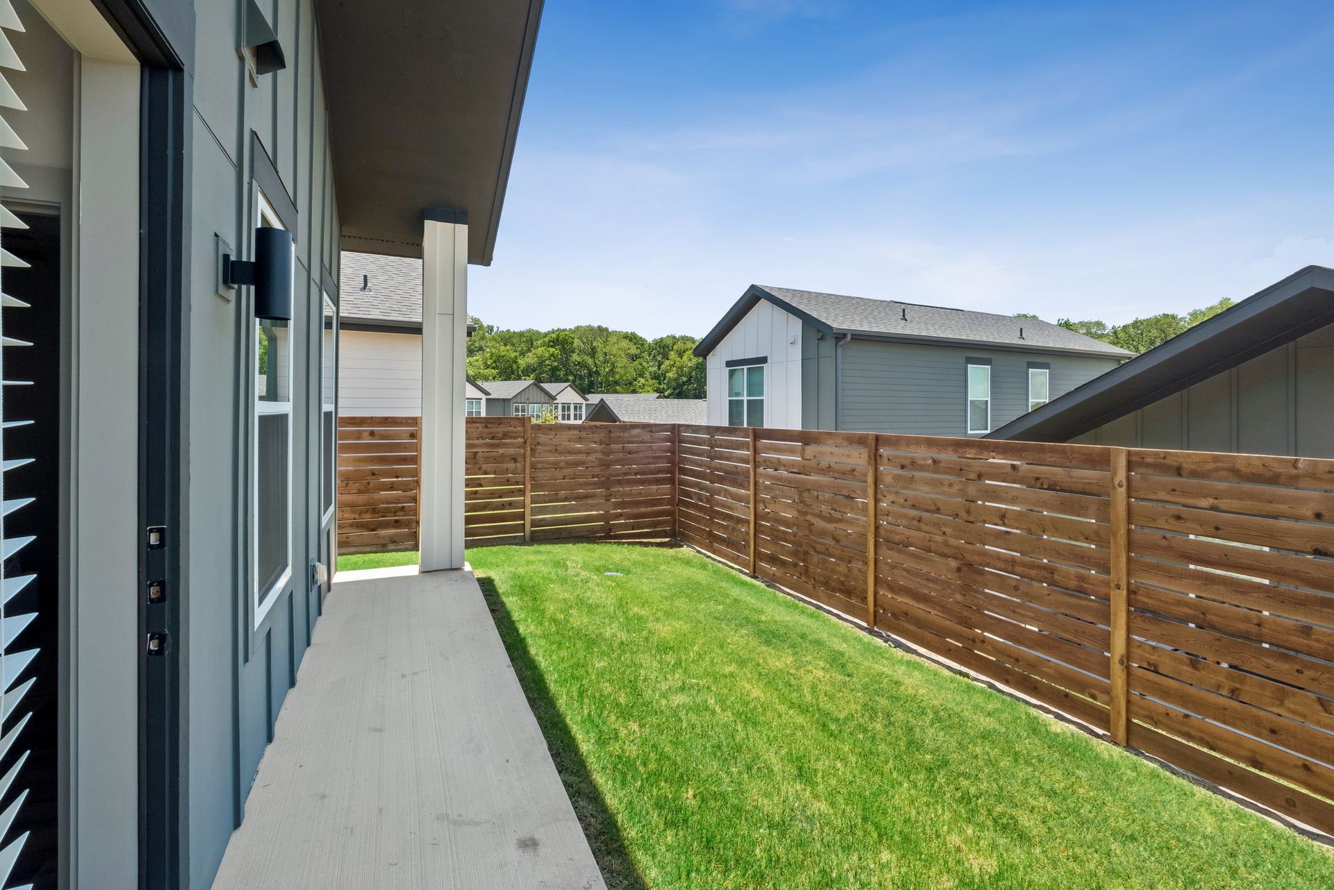 Narrow backyard lawn beside a house, with wooden fence and concrete walkway under a clear sky