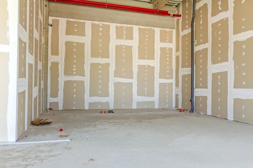 Construcción interior con paneles de yeso parcialmente instalados; paredes color beige con juntas blancas, suelo de hormigón.