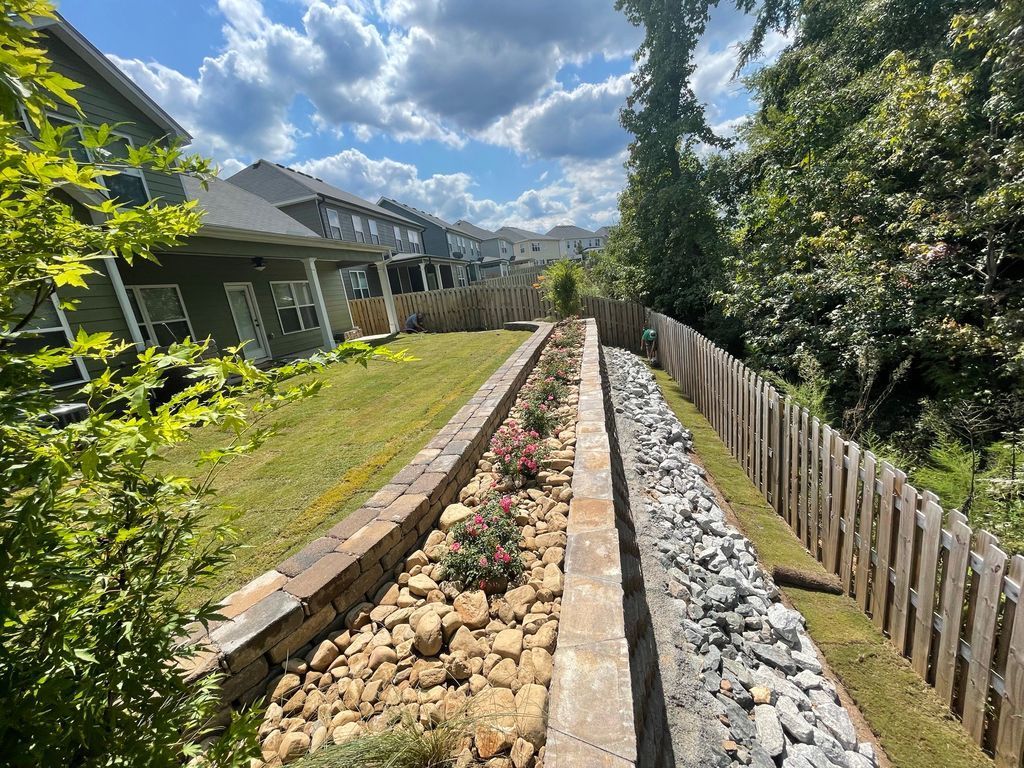 Gravel drainage channel lined with rocks beside homes and a wooden fence under a partly cloudy sky