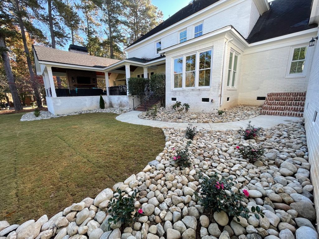 White house with a stone garden bed, lawn, and covered patio in a sunny yard.
