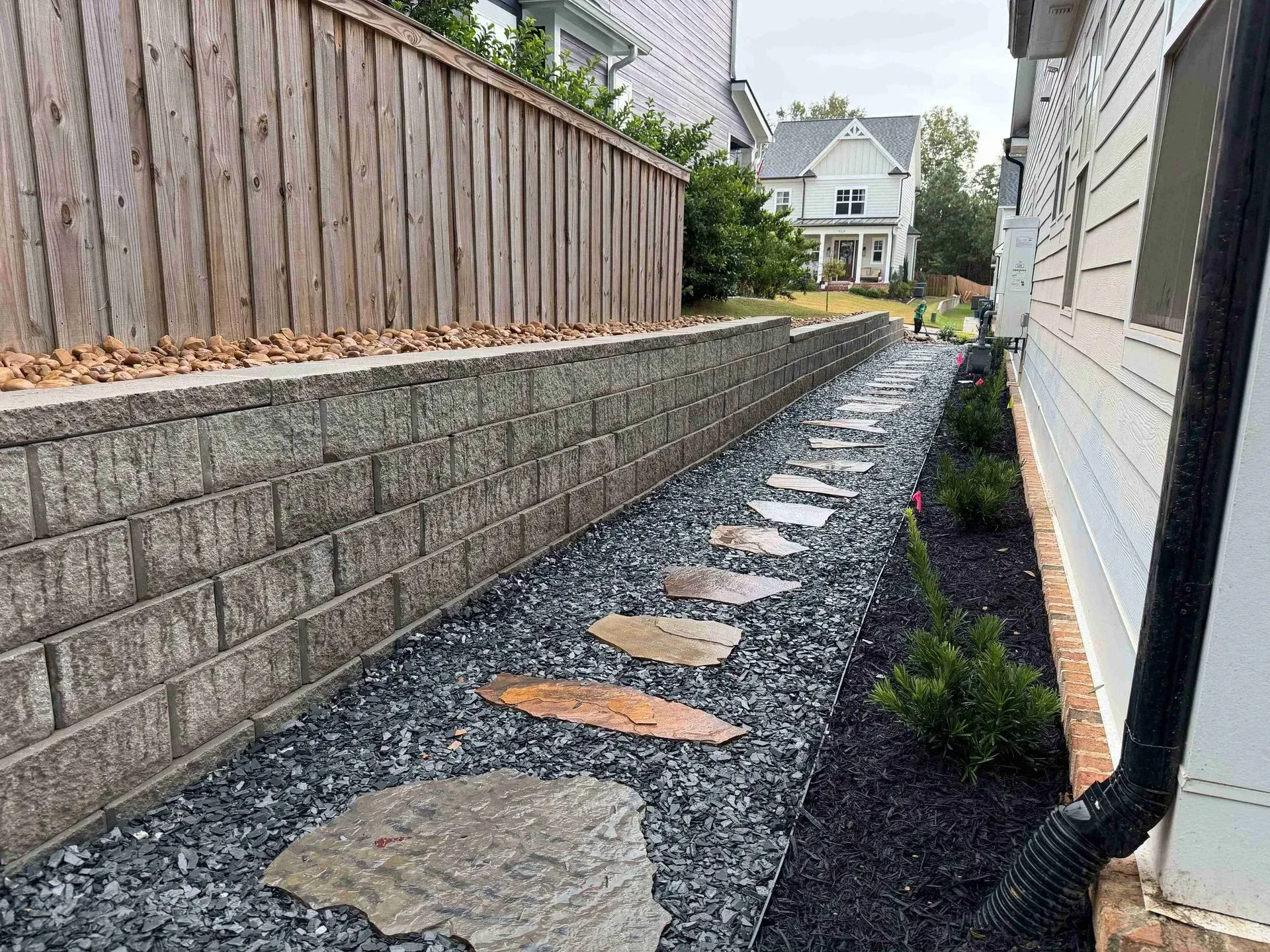 Narrow gravel pathway with stepping stones between a brick wall and house, leading to a backyard.