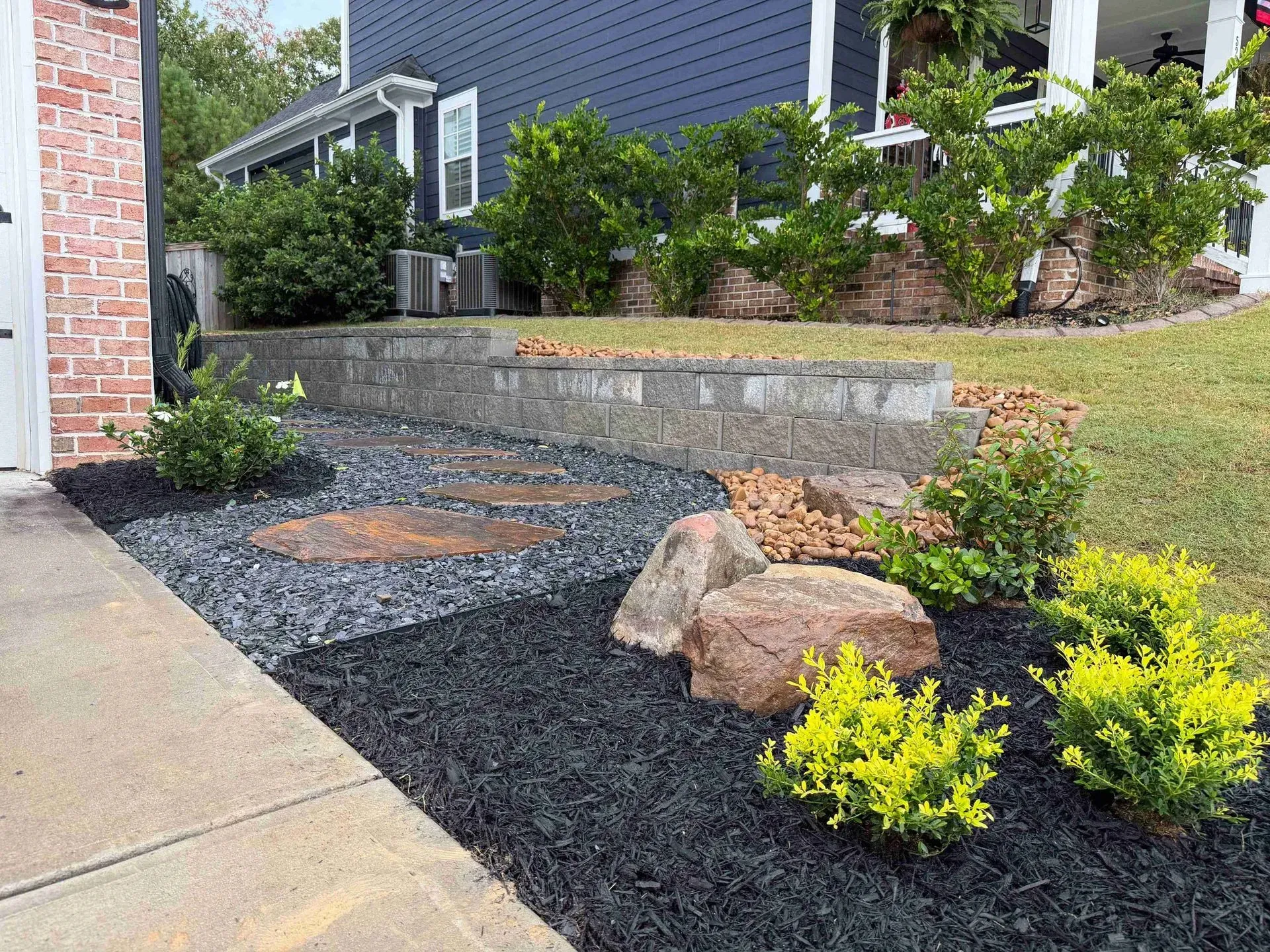 Front yard landscaping with black mulch, rocks, shrubs, and a concrete walkway beside a house