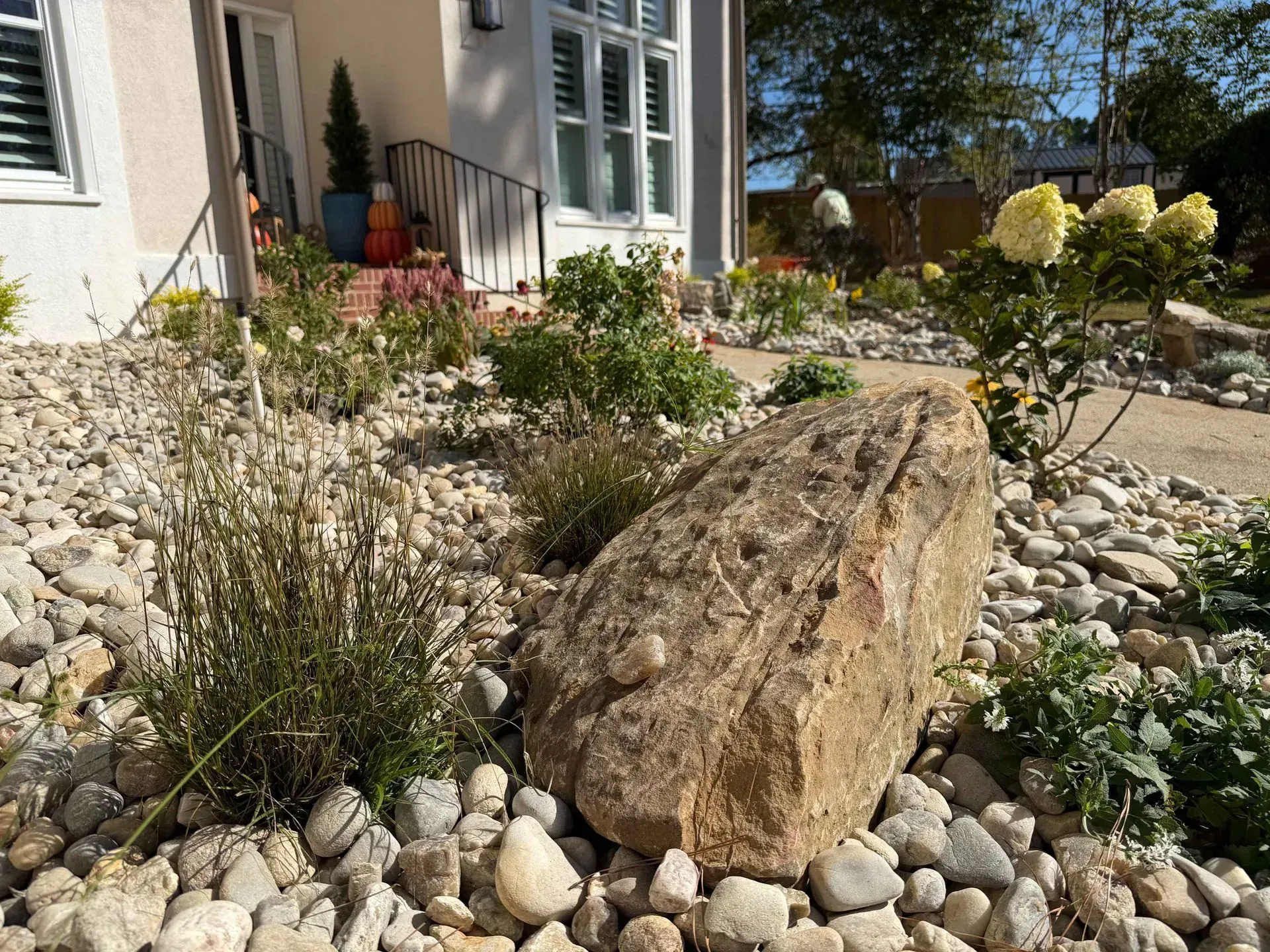 Large boulder in a landscaped front yard with white rocks, shrubs, and a house porch in the background