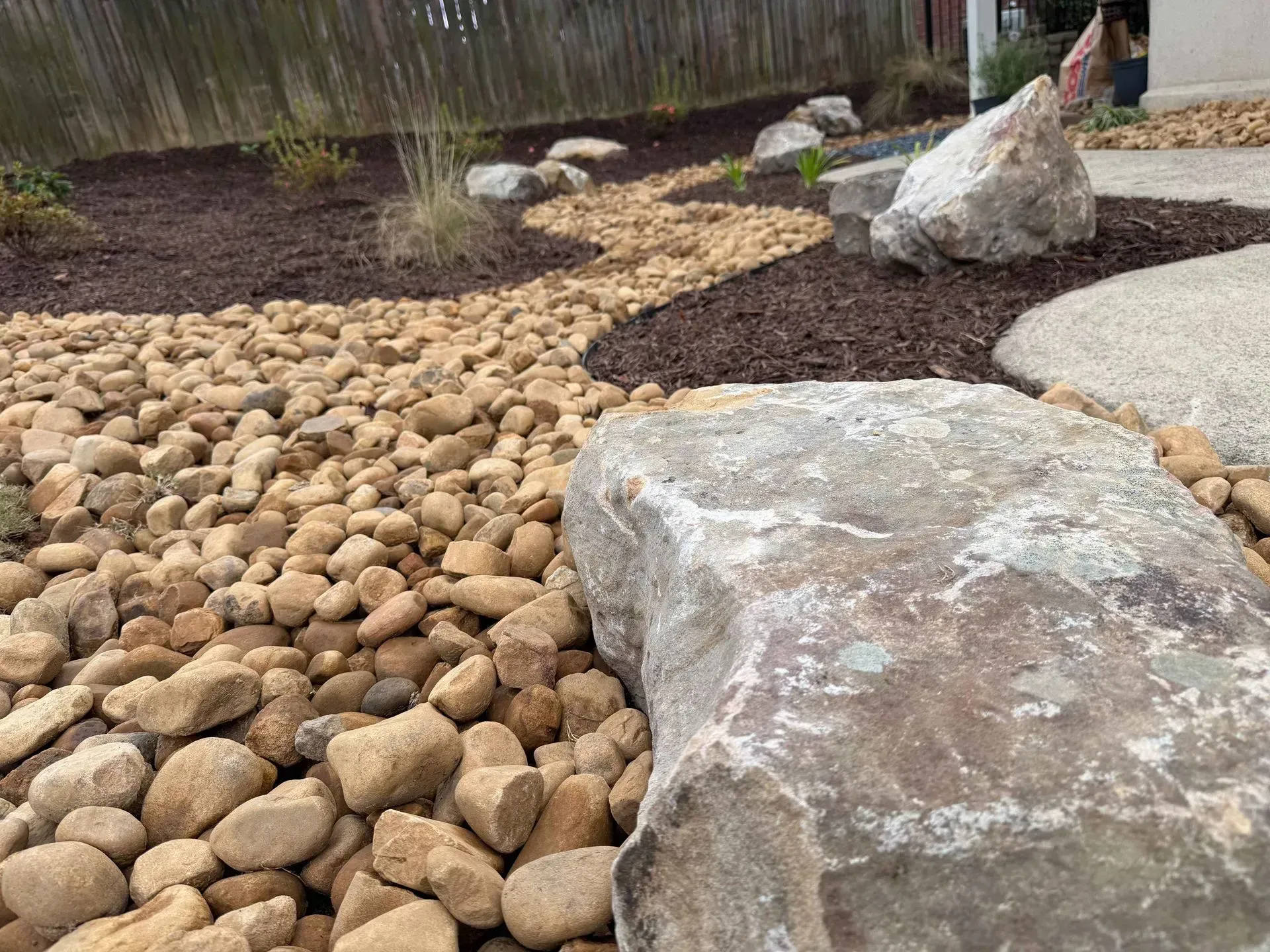 Rock garden with tan pebbles, large gray stones, and dark mulch beside a fence.