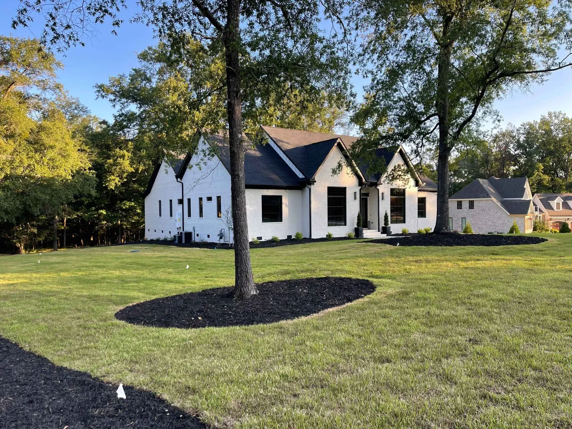 White house with black roof and dark shutters on a grassy lawn with trees and a smaller outbuilding.
