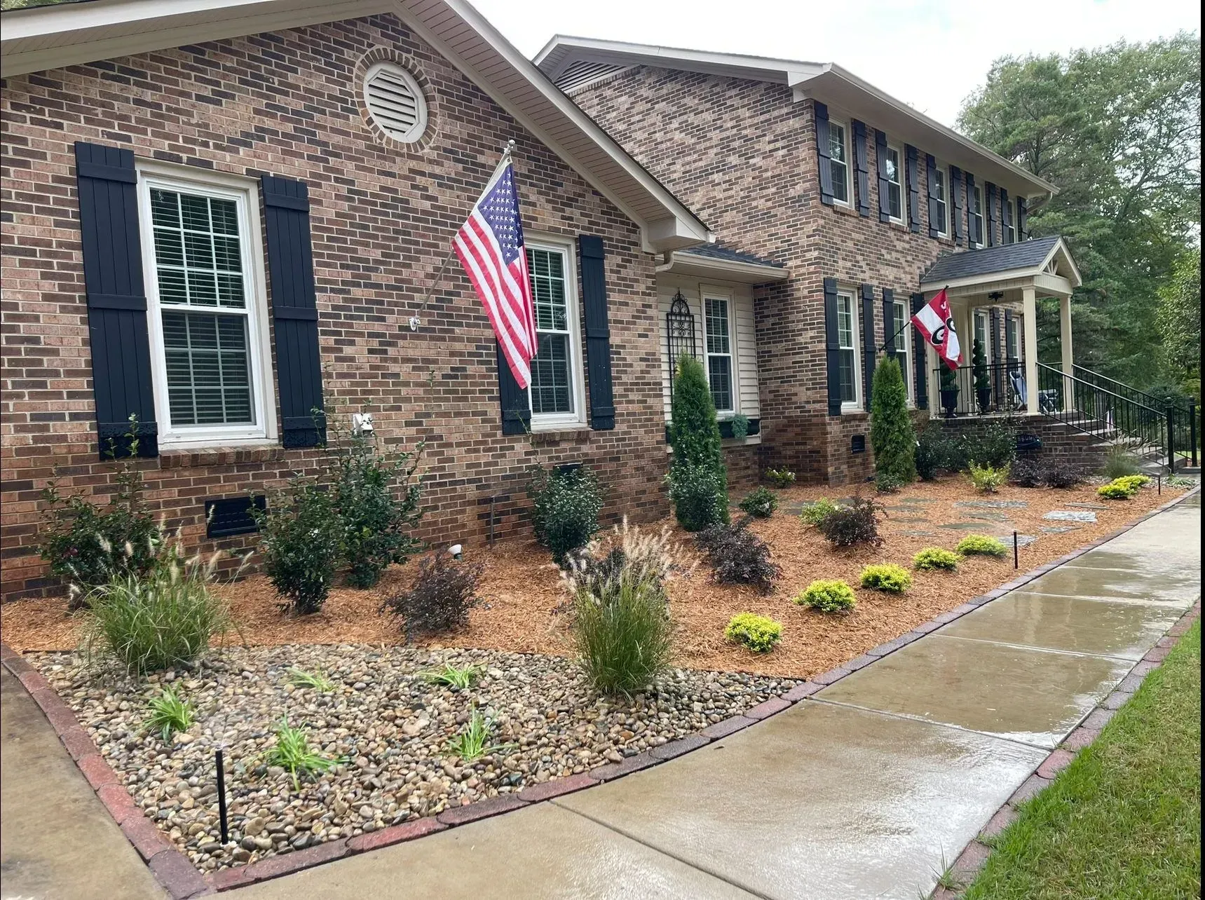 Brick house with black shutters and American flags beside a wet sidewalk and landscaped front yard
