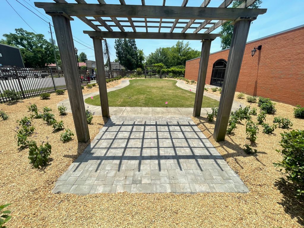 Backyard pergola casting a grid shadow over a stone patio, with shrubs, lawn, and brick walls.