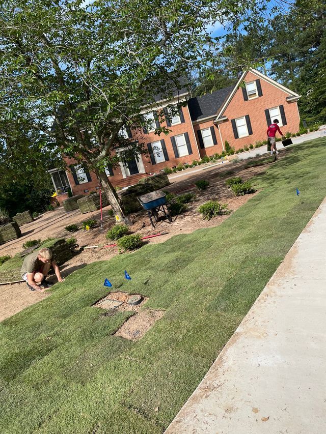 Two people on a grassy campus lawn by brick buildings, one kneeling and one walking in red shirt.