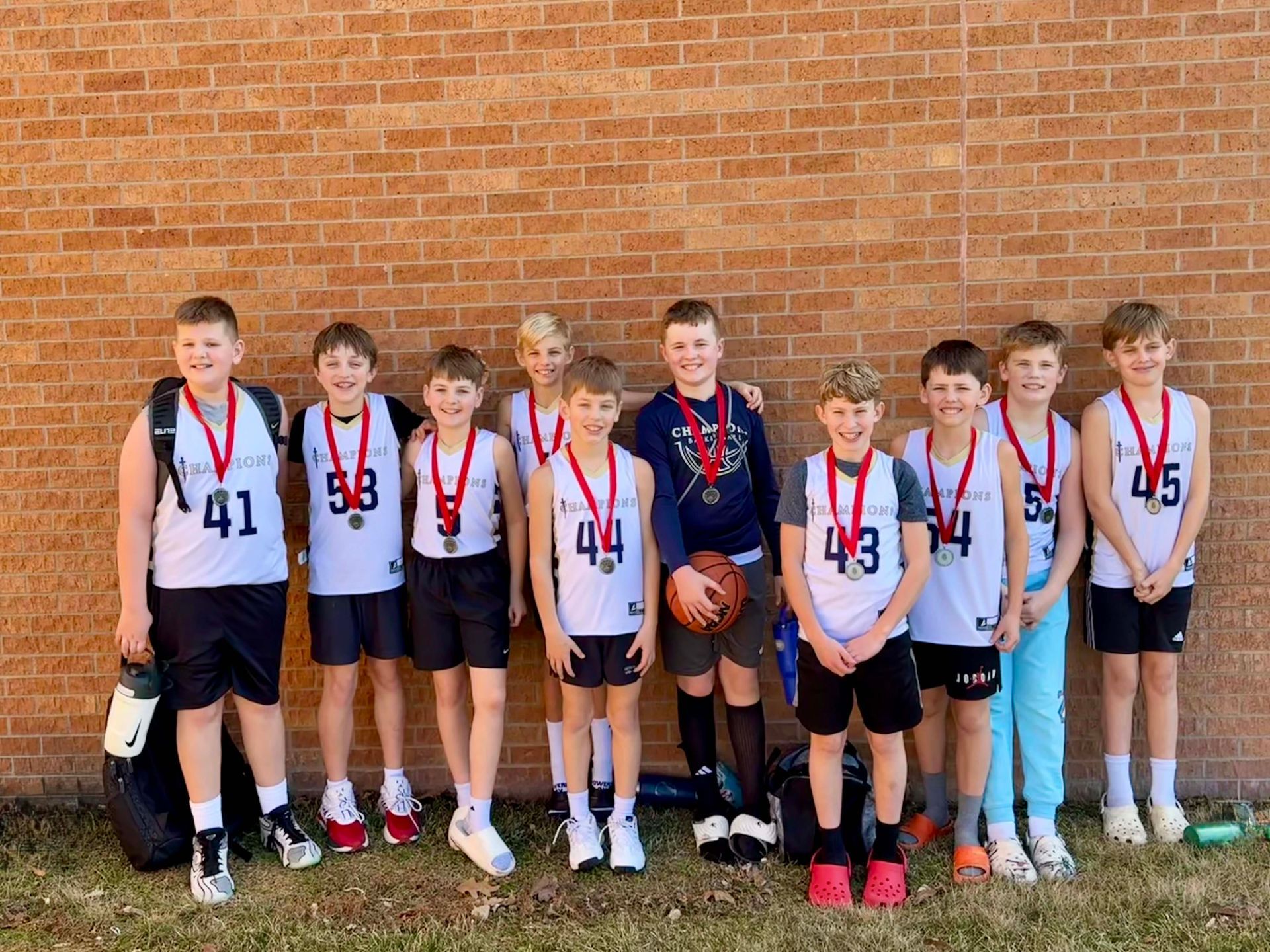 Group of youth basketball players in white jerseys with medals, standing with a basketball in front of a brick wall