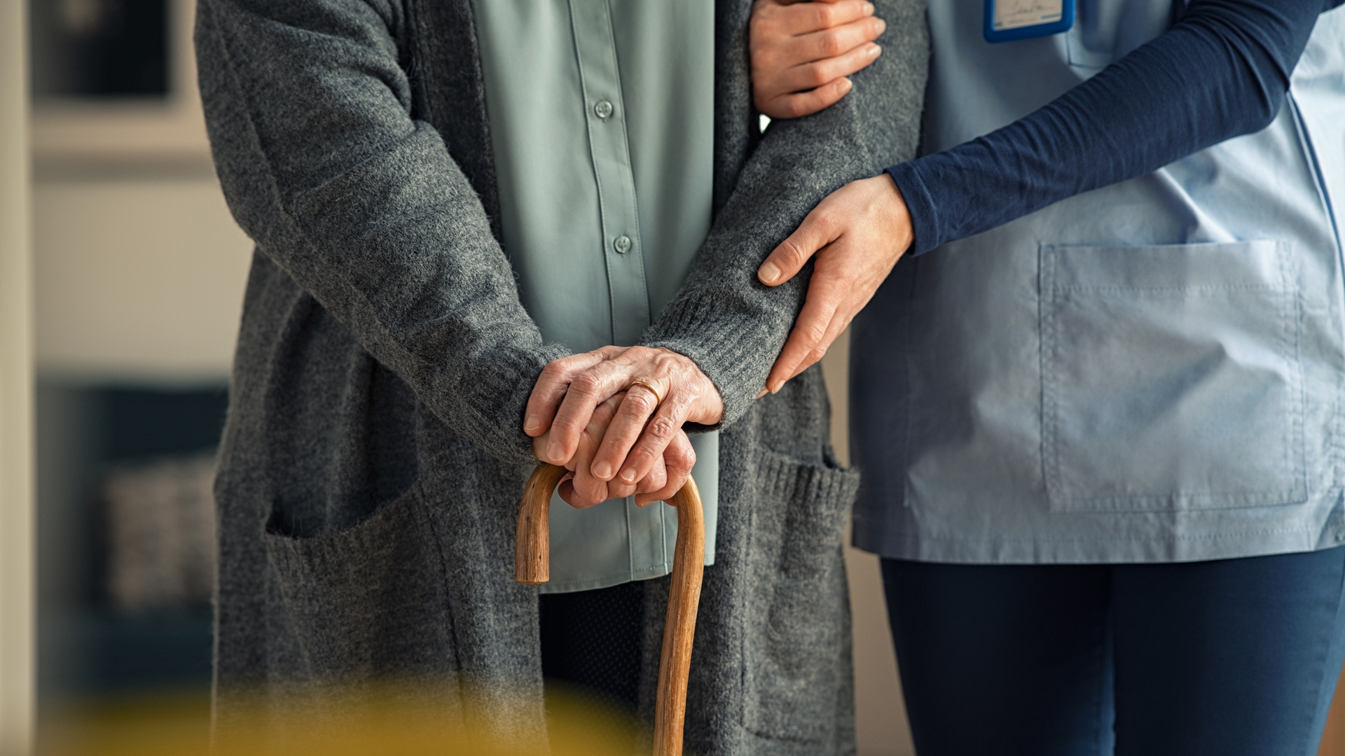 Caretaker supporting an older adult holding a wooden cane, close-up indoors
