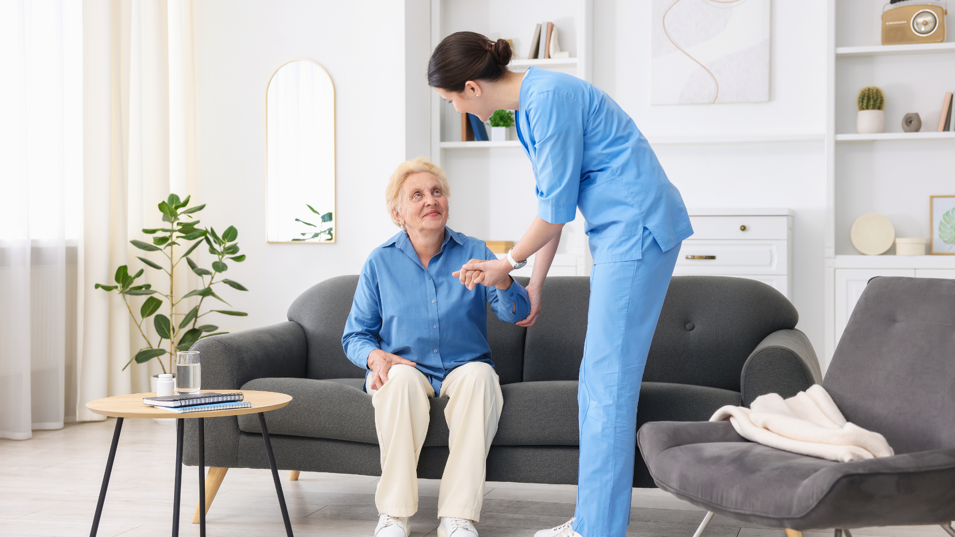 Caregiver in blue scrubs assisting an older adult on a sofa, inside a well-lit living room.