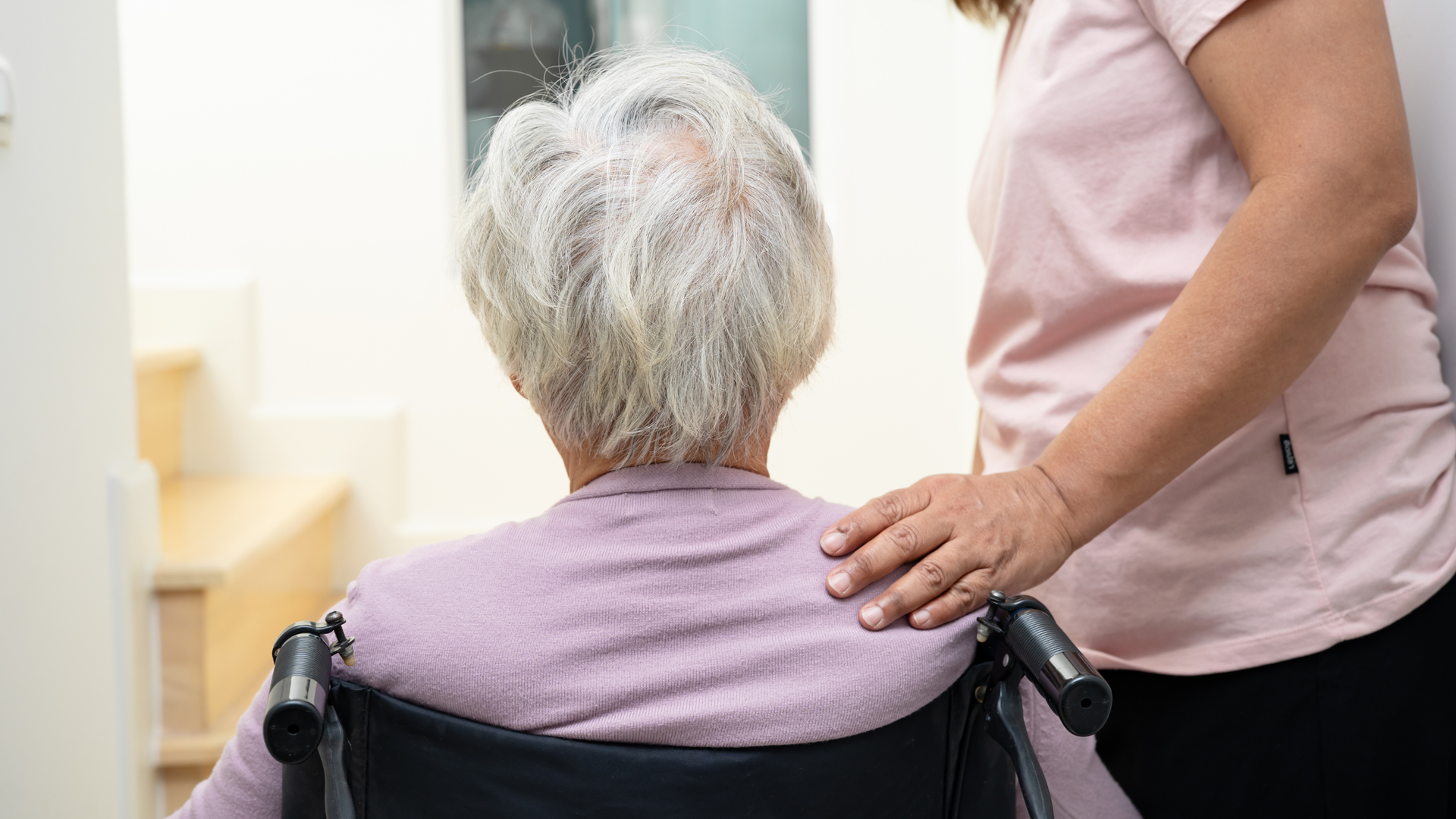 Caregiver comforting a seated person with gray hair, hand on shoulder in a bright room