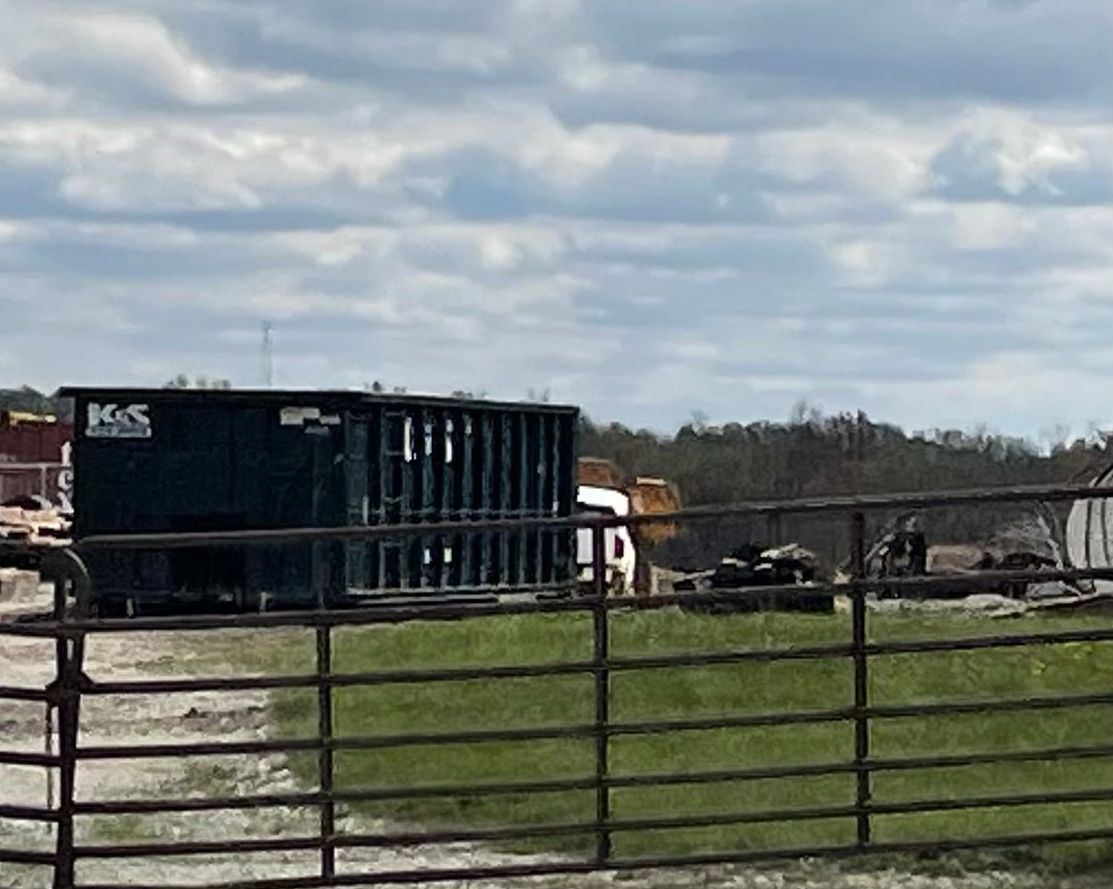A black dumpster is sitting in the middle of a grassy field behind a metal fence.