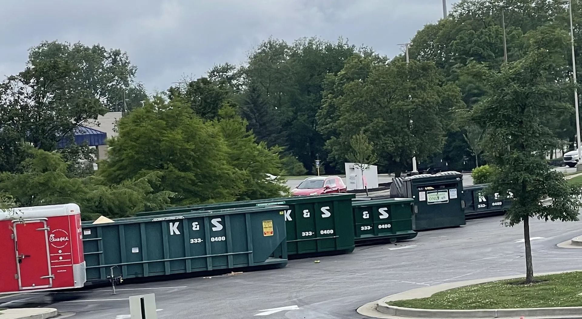 Several large dumpsters line a paved area beside a red portable toilet and trees.