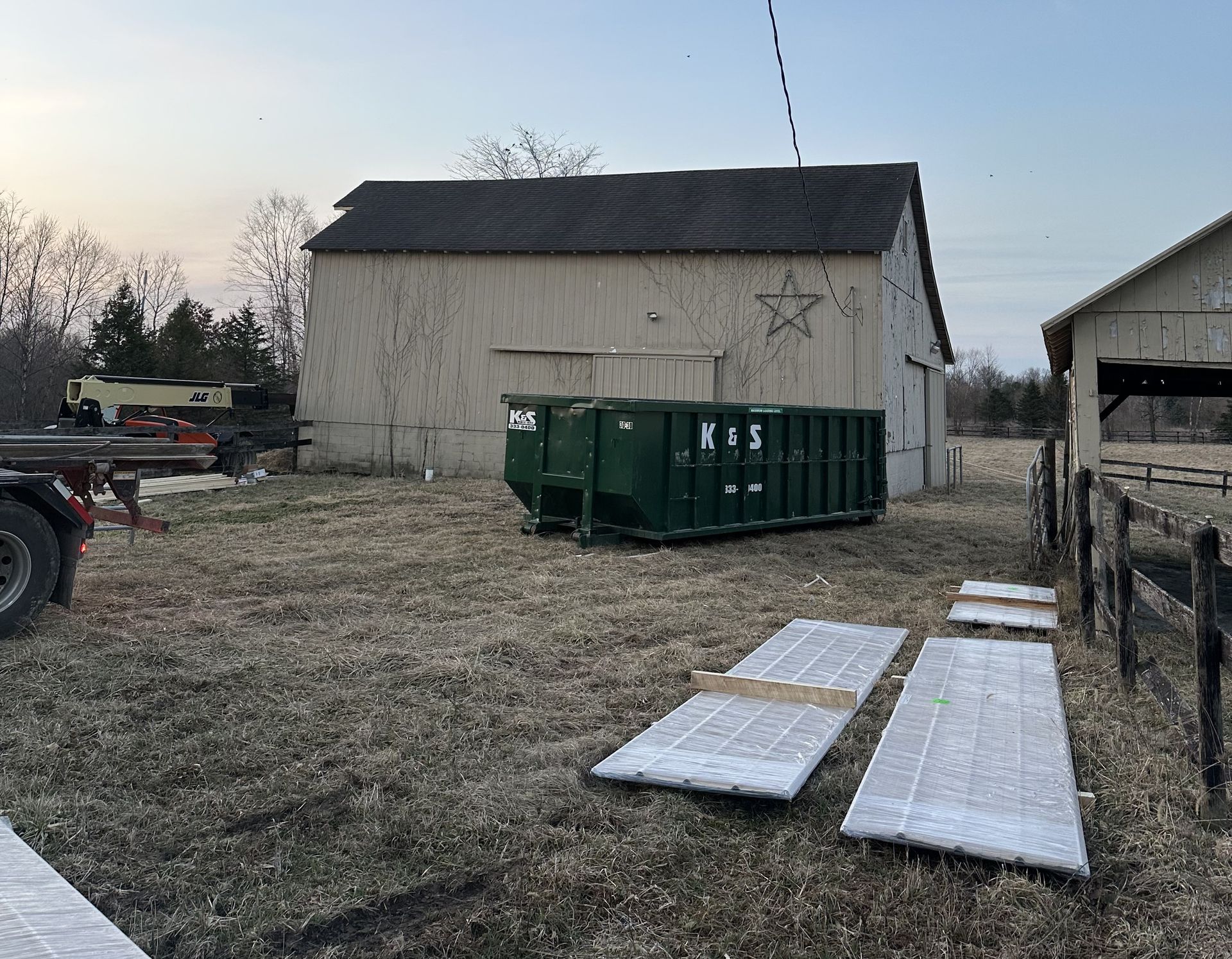 A large green dumpster is sitting in a field next to a barn.