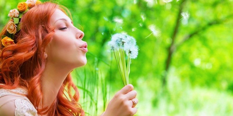 A woman with red hair is blowing a dandelion in a field.