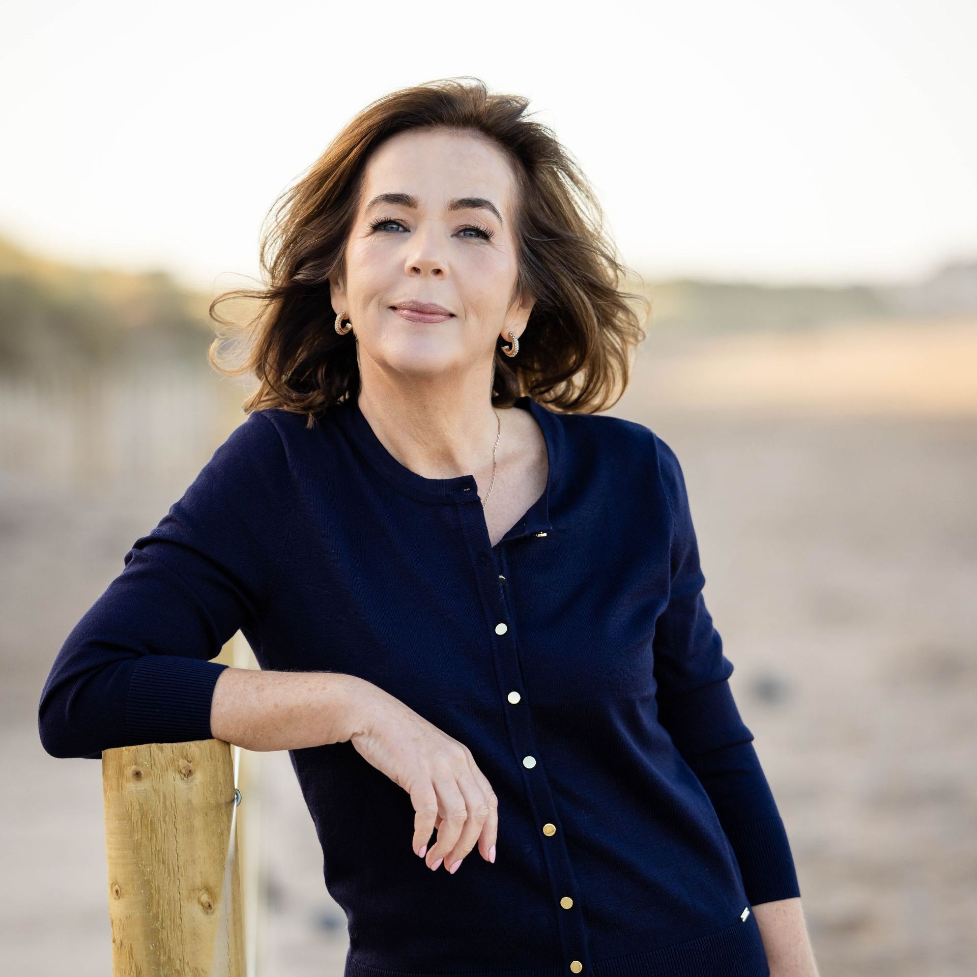 A woman in a blue cardigan leans on a wooden post