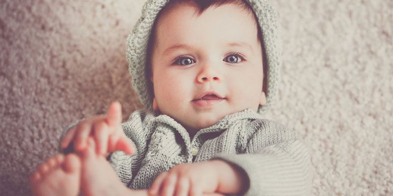 A baby wearing a sweater and hat is laying on a carpet.
