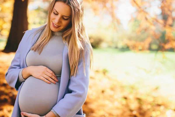 A pregnant woman is standing in a park holding her belly.