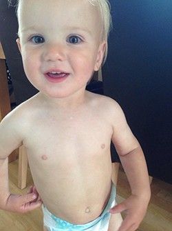 A shirtless baby in a diaper is standing on a wooden chair.