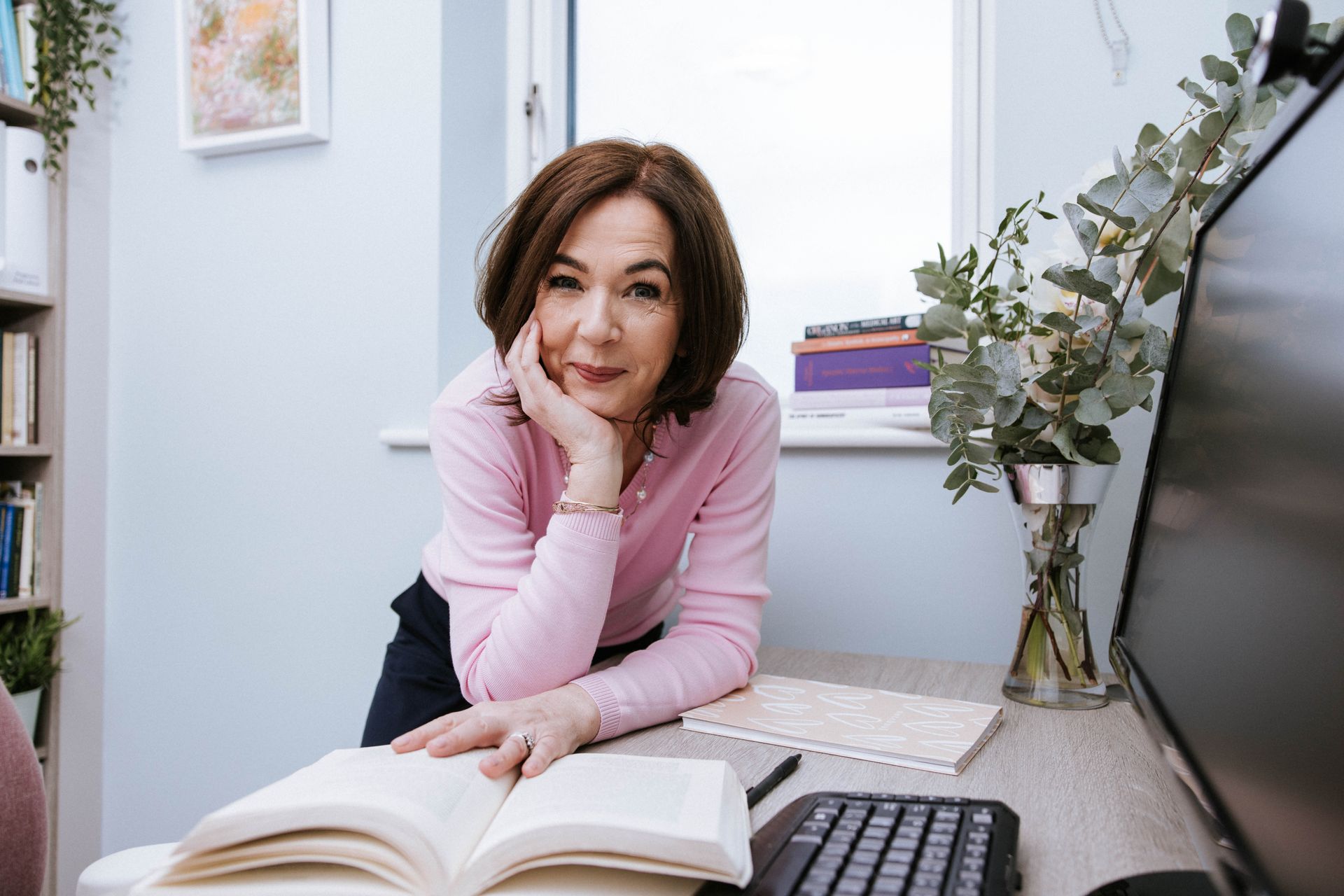 A woman is sitting at a desk with an open book and a computer.