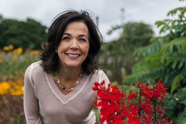 A woman is smiling while standing next to red flowers in a garden.