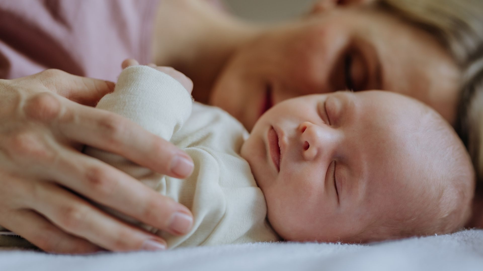 Mother resting with sleeping baby, both on a white bed, soft lighting.